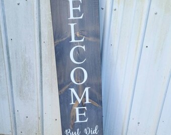 May include: A wooden sign with the word "WELCOME" written vertically in white paint. Below the word is the text "But Did You Text First?" with a small sprig of leaves at the top and bottom of the sign.