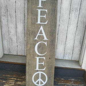 May include: A wooden sign with the word "PEACE" spelled vertically in white letters. The sign has a distressed wood finish and a peace symbol at the bottom.