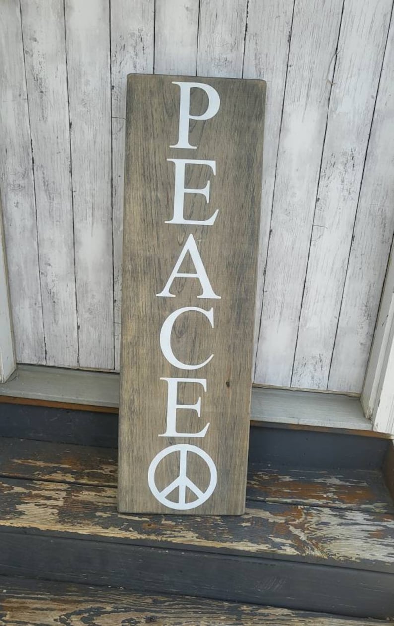 May include: A wooden sign with the word "PEACE" spelled vertically in white letters. The sign has a distressed wood finish and a peace symbol at the bottom.
