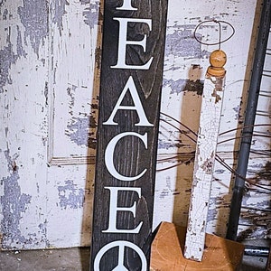 May include: A wooden sign with the word "PEACE" spelled vertically in white letters on a dark brown background. A white peace symbol is at the bottom of the sign.