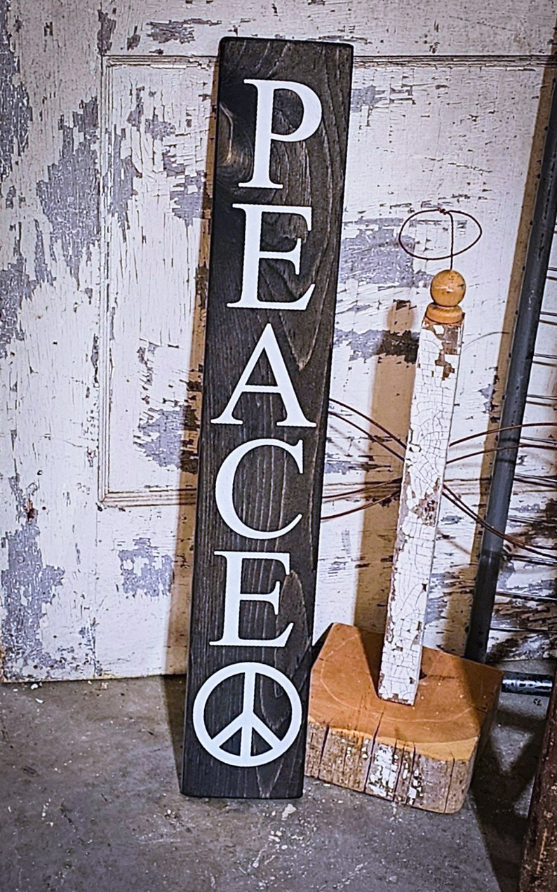 May include: A wooden sign with the word "PEACE" spelled vertically in white letters on a dark brown background. A white peace symbol is at the bottom of the sign.