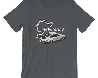 May include: A dark gray t-shirt with a white outline of the Nürburgring race track and a white Porsche 911 car. The text "nürburgring" is printed in brown letters.