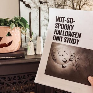 Puede incluir: Un folleto titulado "Not-So-Spooky Halloween Unit Study" con un gráfico de luna llena y murciélagos. La imagen también muestra una maceta con forma de calabaza, velas y decoraciones de murciélagos, creando un tema de Halloween.
