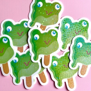 May include: A set of eight green frog popsicle stickers with glitter accents. The stickers are shaped like frogs with googly eyes and a pink tongue.