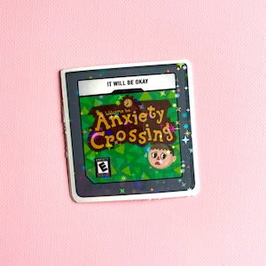 May include: A video game cartridge with the title "Welcome to Anxiety Crossing" and the text "It Will Be Okay." The cartridge has a green and brown background with a cartoon character. The cartridge is on a pink surface.
