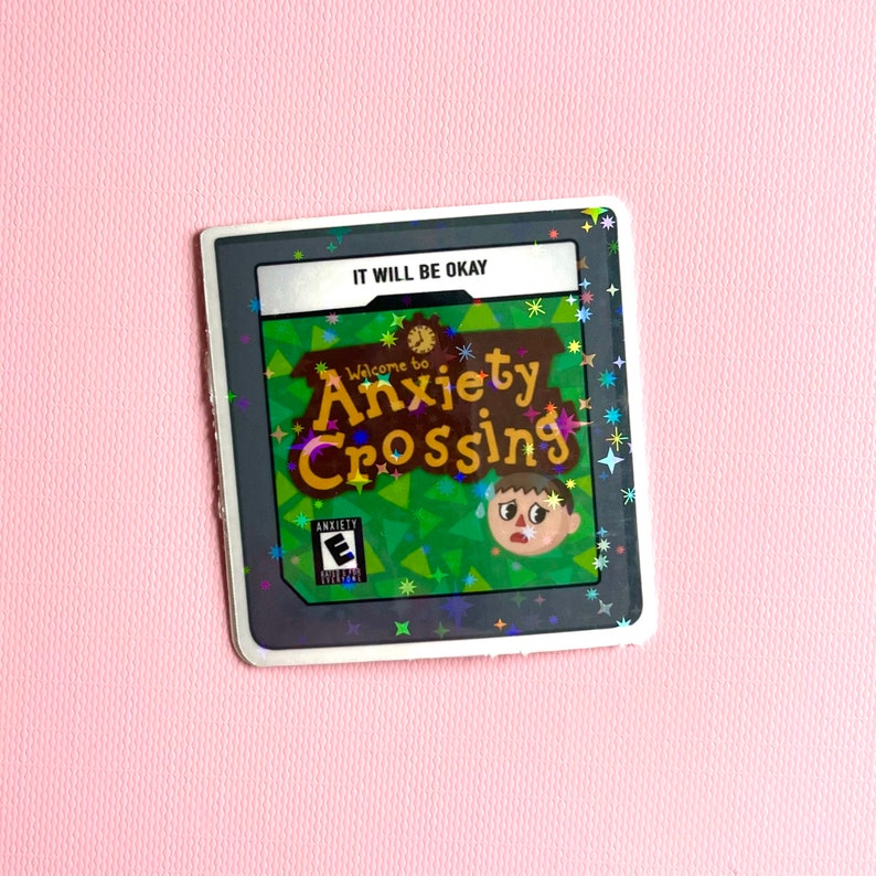 May include: A video game cartridge with the title "Welcome to Anxiety Crossing" and the text "It Will Be Okay." The cartridge has a green and brown background with a cartoon character. The cartridge is on a pink surface.