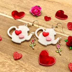 Pode incluir: Brincos de bule brancos com detalhes de coração vermelho e rosa e ganchos dourados. Cada brinco apresenta um pingente pendurado de uma rosa. Espalhadas estão decorações vermelhas em forma de coração e rosas cor-de-rosa, tudo em uma superfície de madeira.
