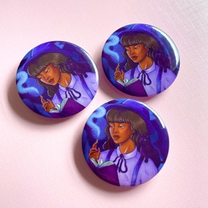 May include: Three round purple and blue buttons with a black cat and a woman wearing a purple hat and a white shirt reading a book. The woman has brown skin and brown hair. The background is a dark blue with white swirls.