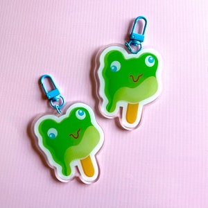 May include: Two green frog popsicle keychains with blue swivel clips. The frog popsicles have a smiling face with big eyes and a stick.