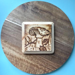 May include: A square wooden plaque with a pyrography mushroom design. The mushroom is detailed with spots and sits among grass and leaves. The plaque is set on a round wooden surface with a blue background.