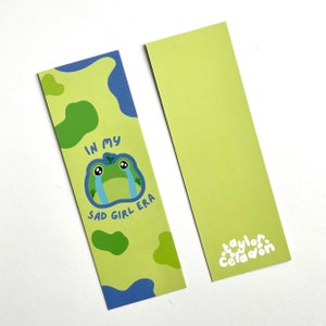 Op de afbeelding: Een groene en blauwe bladwijzer met een huilende cartoonkikker. De bladwijzer heeft de tekst "In my sad girl era" erop. De bladwijzer ligt op een witte achtergrond.