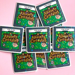 May include: A collection of square stickers featuring the text "Welcome to Anxiety Crossing" with a distressed character design. The stickers have a green and brown color scheme with the text "IT WILL BE OKAY" at the top. The stickers are scattered on a pink surface.