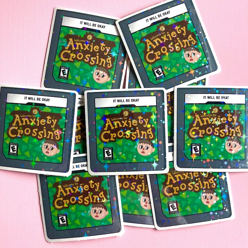 May include: A collection of square stickers featuring the text "Welcome to Anxiety Crossing" with a distressed character design. The stickers have a green and brown color scheme with the text "IT WILL BE OKAY" at the top. The stickers are scattered on a pink surface.
