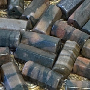 May include: Close-up of a collection of hexagonal beads. The beads are a mix of dark gray, brown, and reddish-brown colors, with a polished, slightly reflective surface. Each bead has a hole drilled through its center.
