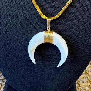 May include: A gold-toned necklace with a crescent-shaped pendant. The pendant is white with gold wire wrapping. The necklace is displayed on a black surface.