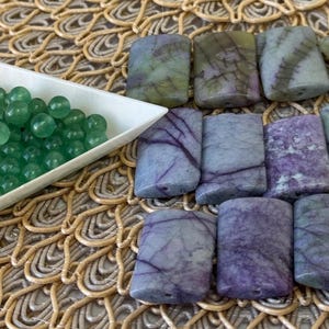 May include: A collection of rectangular, polished stone beads in shades of purple, green, and grey with dark veining, alongside a white triangular tray filled with small, round, translucent green beads, all arranged for jewelry making.