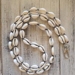 May include: A lanyard featuring a series of white cowrie shells strung along a dark cord. The shells are spaced out with knots between each one. A silver-toned metal lobster clasp is attached to one end of the cord to secure items.