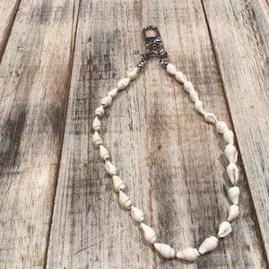 May include: A necklace made of small, white seashells strung together. The necklace is attached to a silver-colored clasp. The seashells are a natural, off-white color and have a smooth texture. The necklace is displayed on a weathered wooden surface.