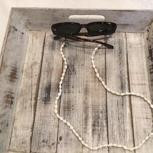 May include: Sunglasses with a tortoiseshell frame and dark lenses rest on a distressed wooden tray. A white seashell eyeglass chain hangs below the glasses, adding a decorative touch. The tray has a white-washed finish.