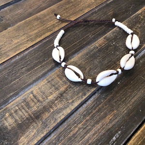 May include: A white shell necklace with brown string and white beads. The necklace is on a wooden surface.