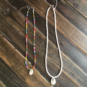 May include: Two beaded necklaces with shell pendants. The necklace on the left has colorful beads and the necklace on the right has white beads.