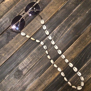 May include: A white shell beaded chain for sunglasses. The chain is made of small, white shells strung together on a black cord. The chain is lying on a wooden surface.