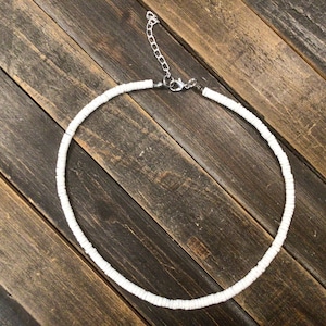Peut inclure: Un collier de perles blanches avec un fermoir argenté. Le collier est composé de petites perles rondes et est posé sur une surface en bois.