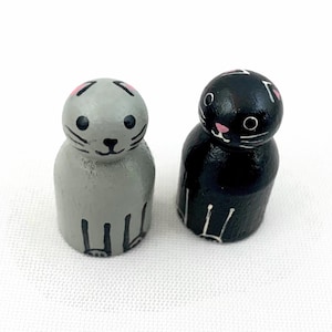 May include: Two wooden cat figurines, one grey and one black, with painted details. The cats have pink ears, black eyes, and white whiskers.