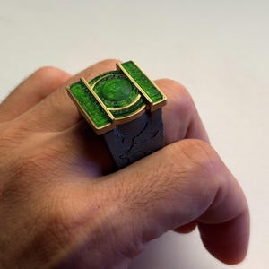 Pode incluir: Um anel com uma banda cinzenta escura texturizada e uma peça superior retangular proeminente. A peça superior é emoldurada em metal dourado e contém um emblema circular verde brilhante e translúcido com detalhes concêntricos no centro.