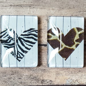 May include: Two glass coasters with a white woodgrain background and a heart-shaped design. One coaster features a zebra print heart and the other features a giraffe print heart.