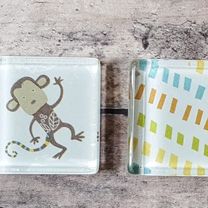 Puede incluir: Dos posavasos cuadrados de vidrio con diseños diferentes. Un posavasos presenta un mono de dibujos animados con un cuerpo marrón y una cola amarilla. El otro posavasos tiene un fondo blanco con un patrón colorido de rayas y rectángulos.