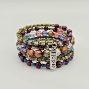May include: A multi-strand wrap bracelet with a variety of beads in shades of purple, gold, blue, and pink. The bracelet features a silver charm that reads "Hope". The beads are of different shapes and sizes.