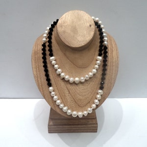 May include: Two necklaces displayed on a wooden bust. One necklace features white pearls, and the other is composed of black beads. The necklaces are draped to showcase their design and length.