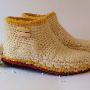 One crocheted slippers with a white base, yellow trim, and red accents. The slippers have a button detail on the top.