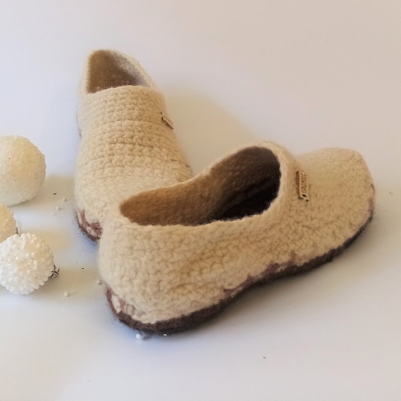 A pairs of beige crocheted slippers with brown soles. The slippers have a label that reads 'Handmade'.