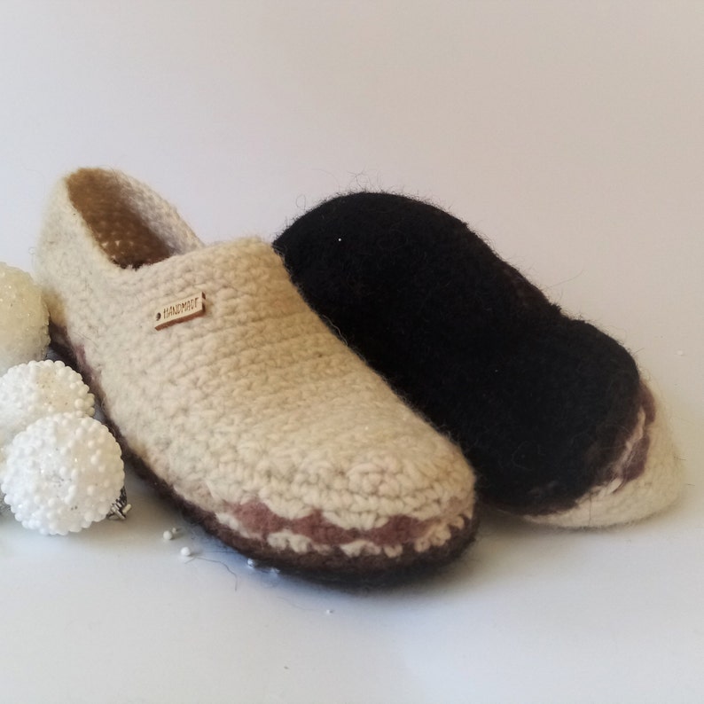 One pairs of crocheted wool slippers. Pair is beige with brown trim with black sole. The beige slippers have a wooden tag that says 'Handmade'.