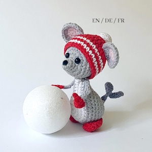 May include: A crocheted gray mouse wearing a red and white striped hat, red mittens, and red shoes, holding a white ball. The mouse has black eyes and a gray tail. The text "EN / DE / FR" is in the top center.