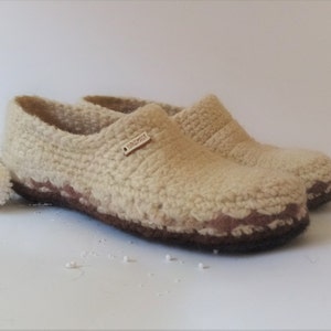One pairs of beige wool slippers with brown soles. The slippers have a crocheted texture and one slipper has a brown leather tag that reads 'HANDMADE'.