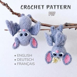 Pode incluir: Dois morcegos de crochê cinzentos pendurados num galho. O texto diz CROCHET PATTERN PDF, ENGLISH, DEUTSCH, FRANÇAIS. Os morcegos têm olhos pretos grandes, orelhas cor-de-rosa e narizes pequenos cor-de-rosa. Um segura uma pequena flor amarela.