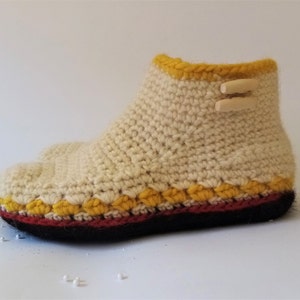 Crocheted cream-colored slipper with a yellow trim and a red and black sole. The slipper has two wooden toggles on the top.