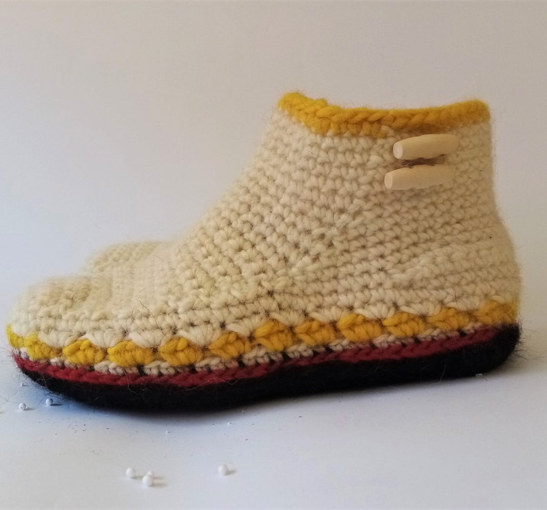 Crocheted cream-colored slipper with a yellow trim and a red and black sole. The slipper has two wooden toggles on the top.
