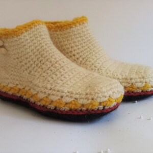 One crocheted slippers in cream with yellow and red trim. The slippers are made of a soft, textured yarn and have a simple design. The slippers are displayed on a white surface.