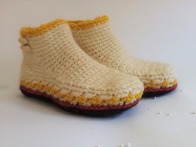 One crocheted slippers in cream with yellow and red trim. The slippers are made of a soft, textured yarn and have a simple design. The slippers are displayed on a white surface.