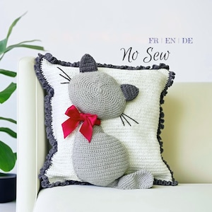 May include: Crocheted cat pillow with a gray cat design, a red bow, and black whiskers. The pillow is white with a gray border and the text "No Sew". This decorative pillow is a charming addition to any room.