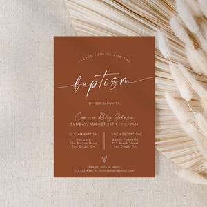 May include: A terracotta-colored invitation card with white script text announcing a baptism. The card includes details such as the date, time, and location of the ceremony and reception. The card is set against a backdrop of dried flowers.