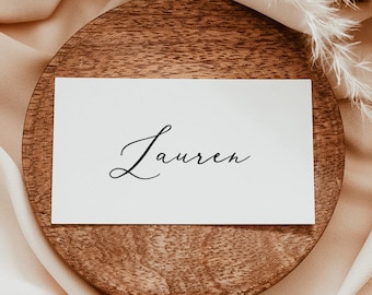 Op de afbeelding: Een witte kaart met de naam "Lauren" in cursief op een houten onderzetter. De onderzetter ligt op een lichtroze stoffen achtergrond met een gedroogde pampasgras tak.