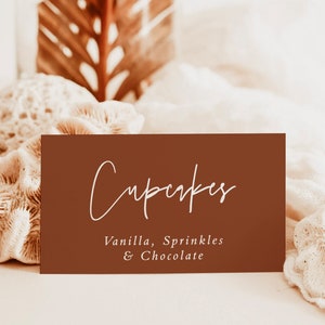 May include: A brown card with white text that reads "Cupcakes" and "Vanilla, Sprinkles & Chocolate". The card is sitting on a white surface with coral and a brown palm leaf in the background.