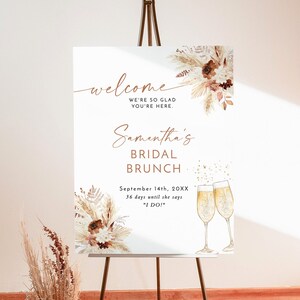 Könnte beinhalten: Ein weißes Schild mit einer Willkommensnachricht für Samanthas Braut-Brunch. Das Schild ist mit getrockneten Blumen und goldenem Glitzer verziert. Der Text lautet: "Willkommen, wir freuen uns, dass Sie da sind. Samanthas Braut-Brunch, 14. September 20XX, 36 Tage bis sie "Ja" sagt!"