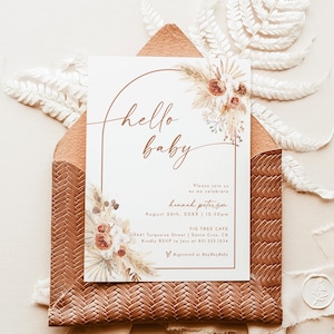 Op de afbeelding: Een uitnodiging voor een babyshower met een aquarelillustratie van gedroogde bloemen en pampasgras. De uitnodiging is gedrukt op een crèmekleurige kaart met een bruine envelop. De tekst op de uitnodiging luidt "hello baby" en bevat de naam "Hannah Peterson", de datum "August 26th, 20XX", de tijd "12:30pm", de locatie "Fig Tree Cafe", het adres "441 Turquoise Street | Santa Cruz, CA" en de RSVP-informatie "Kindly RSVP to Jess at 631.555.1234". De uitnodiging bevat ook de tekst "Registered at Bay Bay Baby".