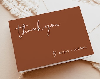 May include: A brown thank you card with white script that says "thank you" and "Avery + Jordan" with a small heart symbol. The card is on a white envelope and a light brown background.